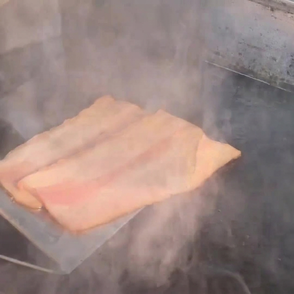 Strisce di bacon che friggono sulla piastra emanando vapore.