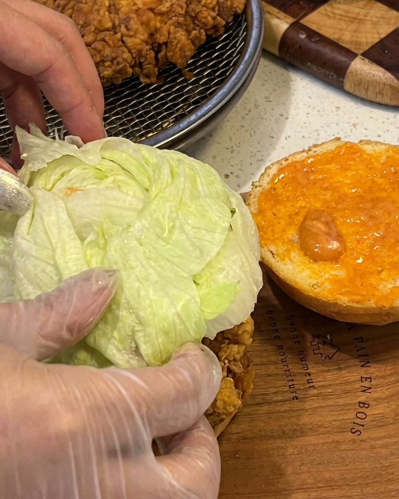 Assemblaggio di un burger di pollo con un filetto croccante, lattuga fresca e salsa su un panino tostato.