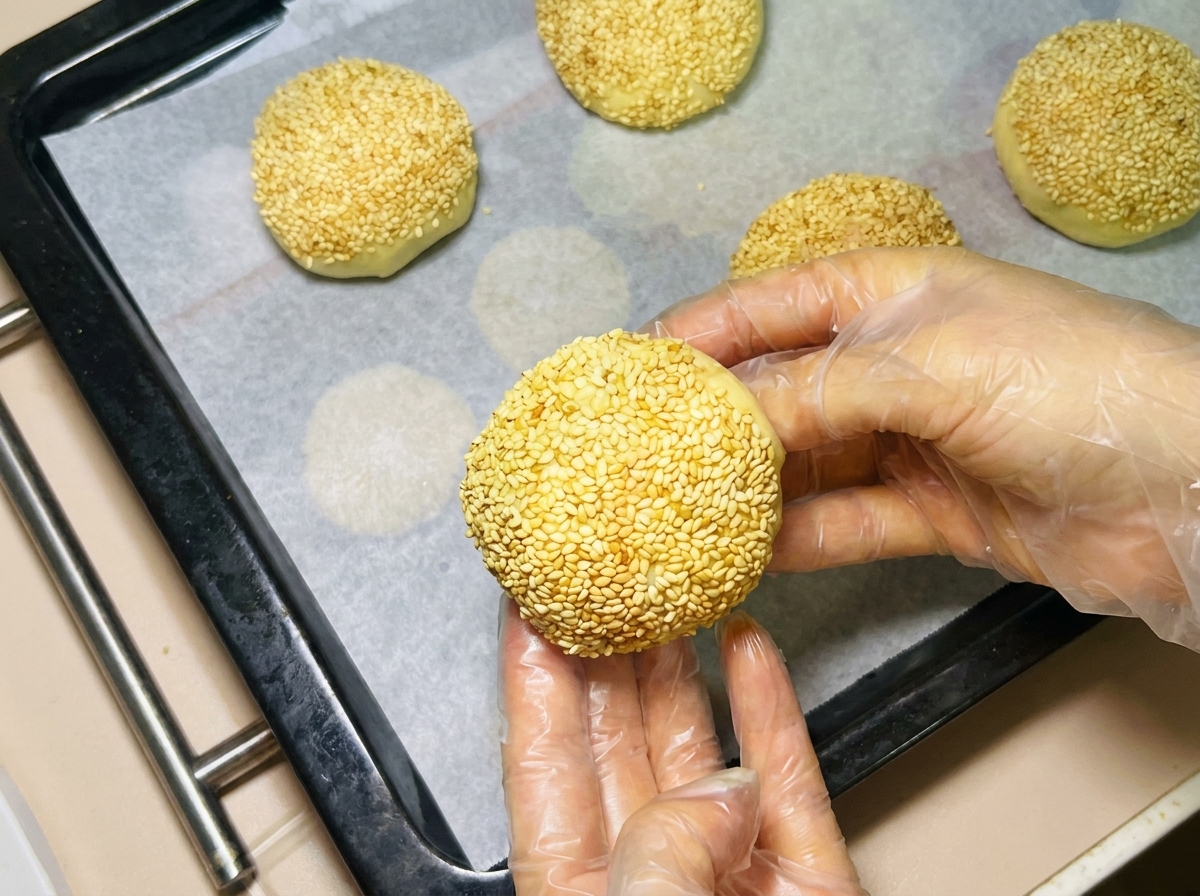Una mano guantata che dispone una pallina di impasto ricoperta di sesamo su una teglia foderata di carta forno.