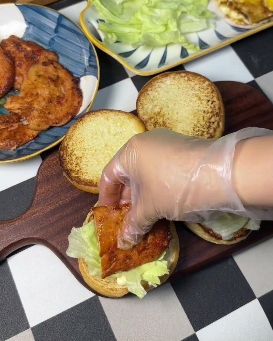 Una mano guantata che posiziona un burger di pollo su un letto di lattuga fresca.