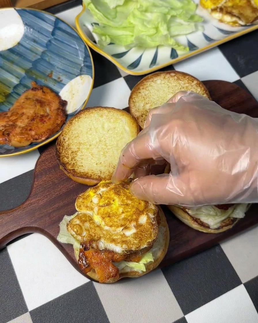 Una mano che posiziona un uovo fritto sopra il burger di pollo.