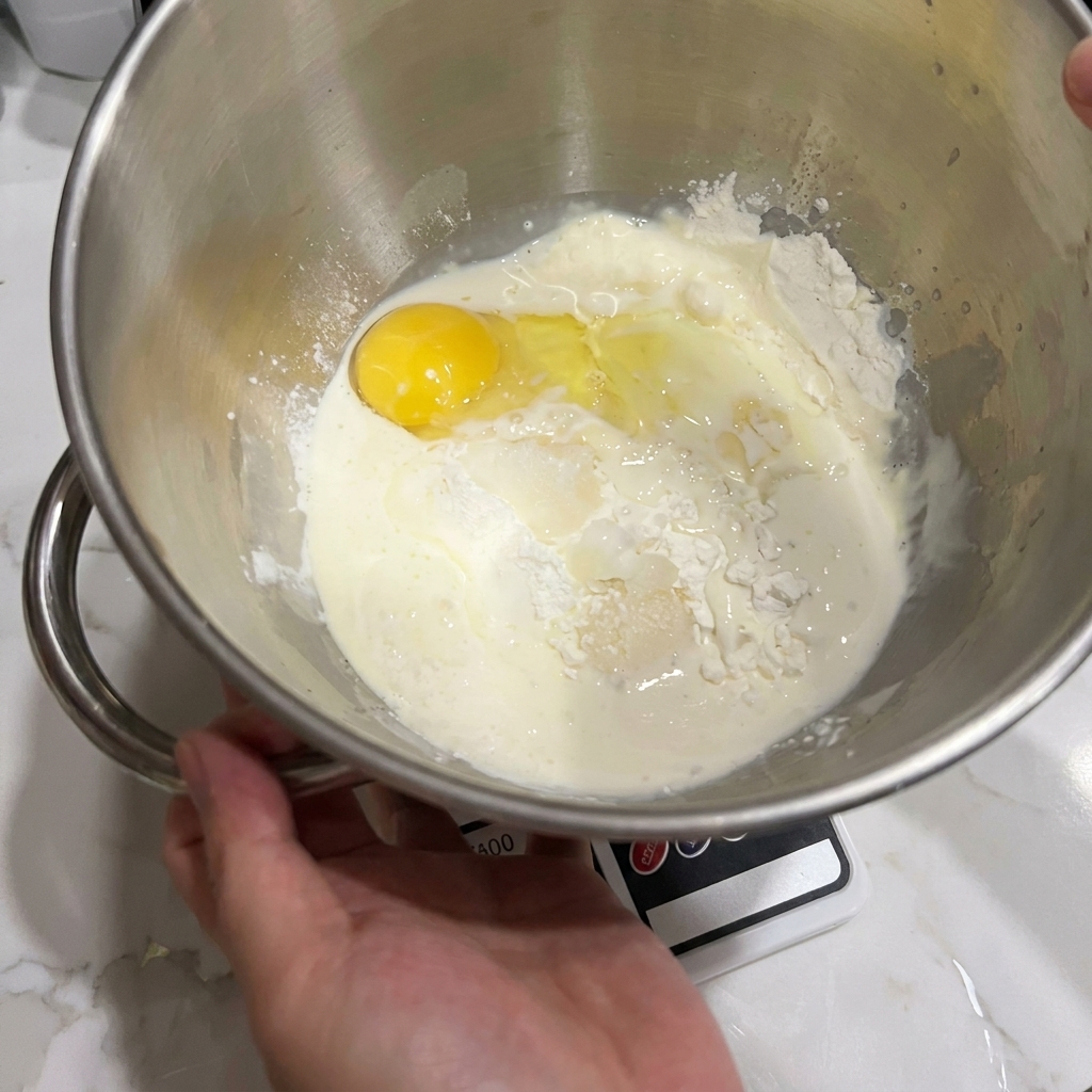 Una ciotola di unimpastatrice con farina, un uovo rotto e latte, pronti per essere mescolati.