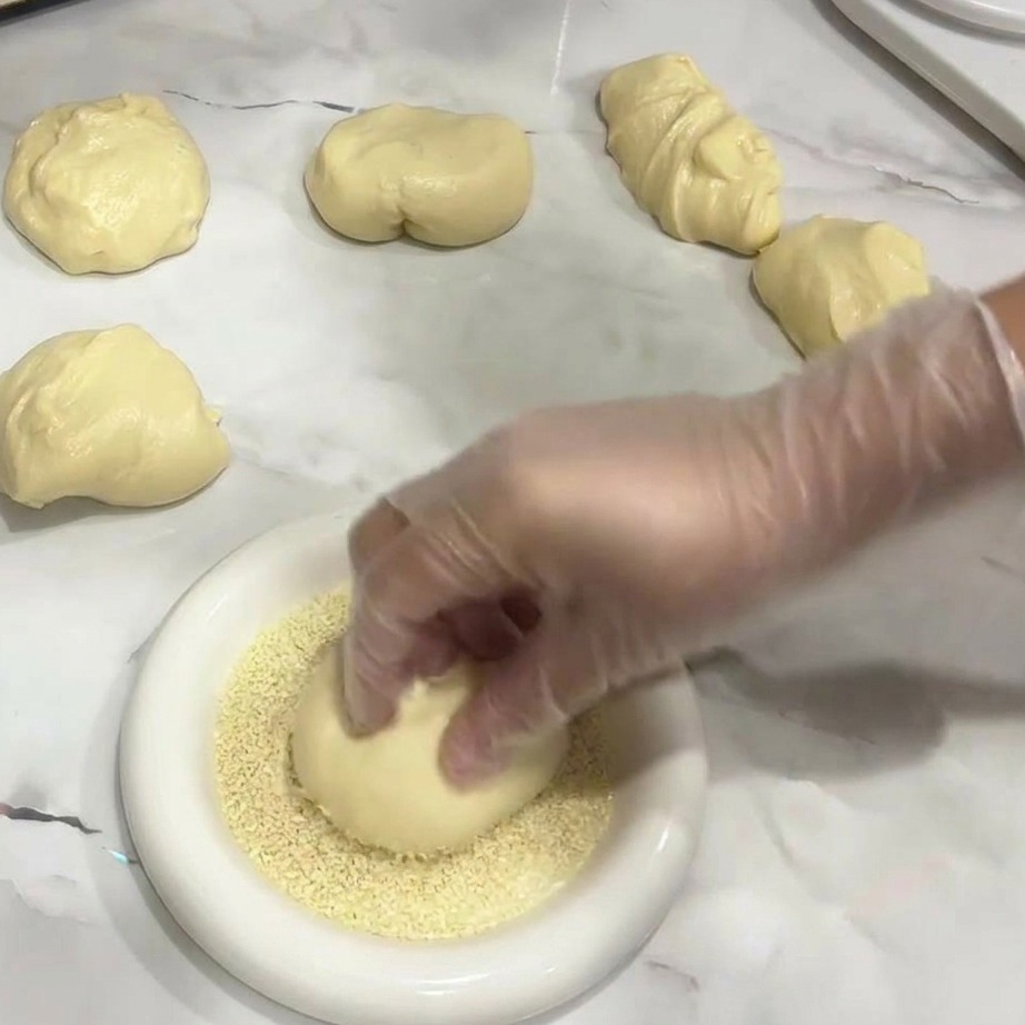 Una mano che posiziona una pallina di impasto liscia e rotonda in un piatto di semi di sesamo bianchi.