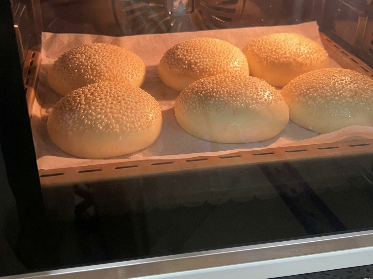 Sei panini per hamburger al sesamo appena sfornati, dorati, seduti su una teglia foderata di carta forno allinterno di un forno.