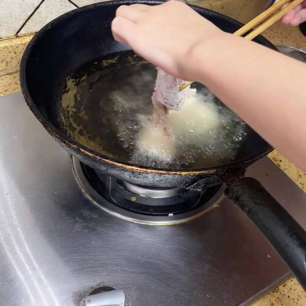 Una persona che usa le bacchette per abbassare delicatamente un pezzo di pollo impanato in un wok nero riempito di olio caldo e gorgogliante.