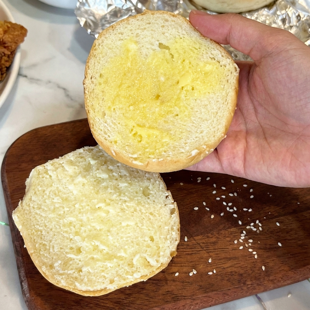 La metà tagliata di un panino al sesamo spalmata di burro fuso, tenuta sopra un tagliere di legno.