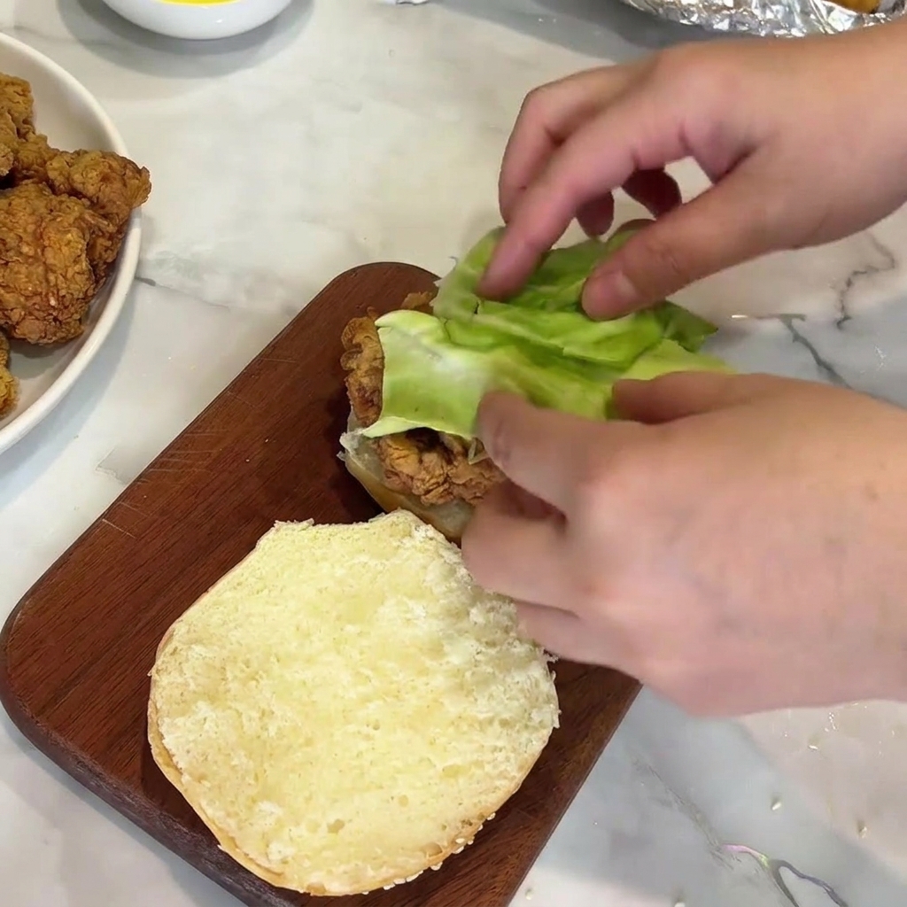 Mani che posizionano una foglia di lattuga verde fresca sopra una cotoletta di pollo croccante su un panino.