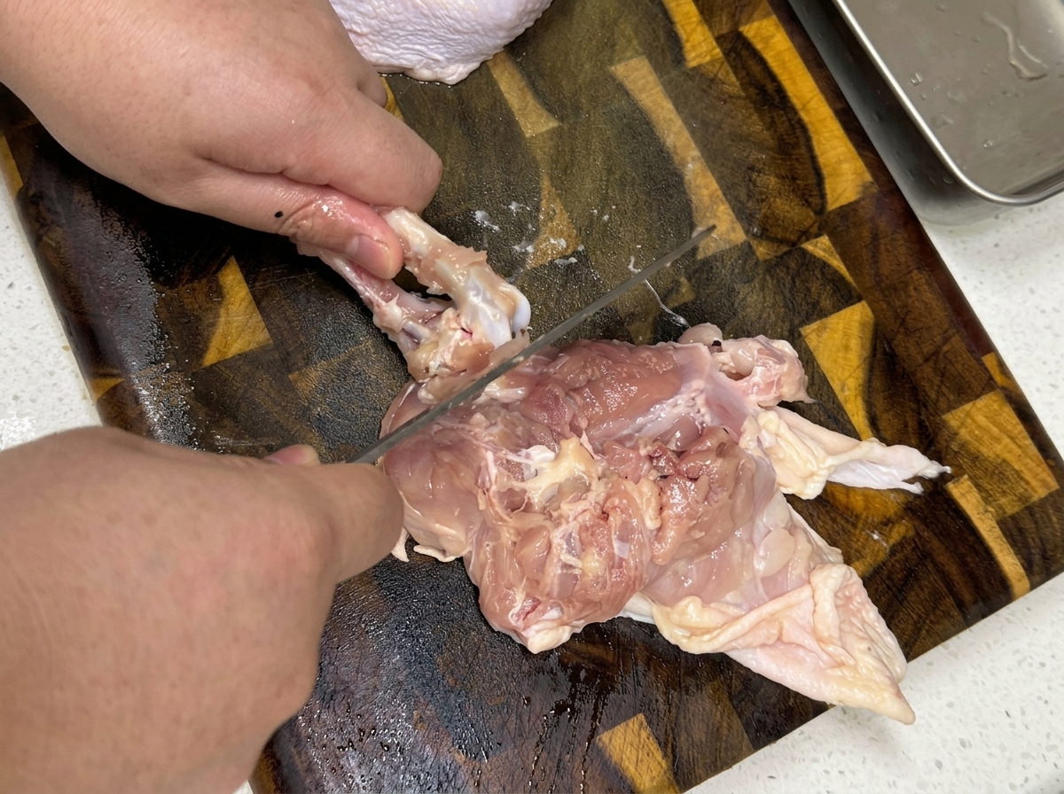 Un primo piano delle mani che disossano una coscia di pollo cruda su un tagliere di legno.