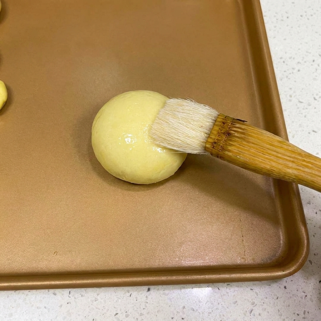 Un pennello da pasticceria che applica un leggero strato dacqua sopra una pallina di impasto arrotondata.
