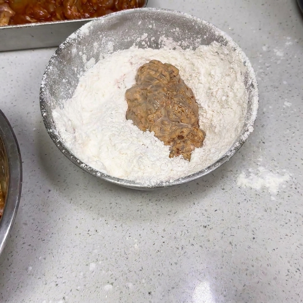 Un pezzo di pollo bagnato e pastellato mescolato in una ciotola dargento piena di farina bianca secca.