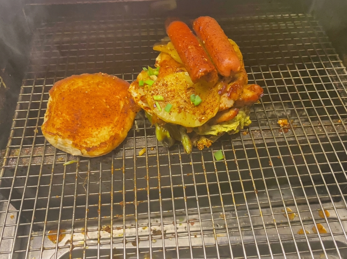 Un burger imponente carico di carni, patate grigliate e salsicce accanto a un panino tostato sulla griglia.