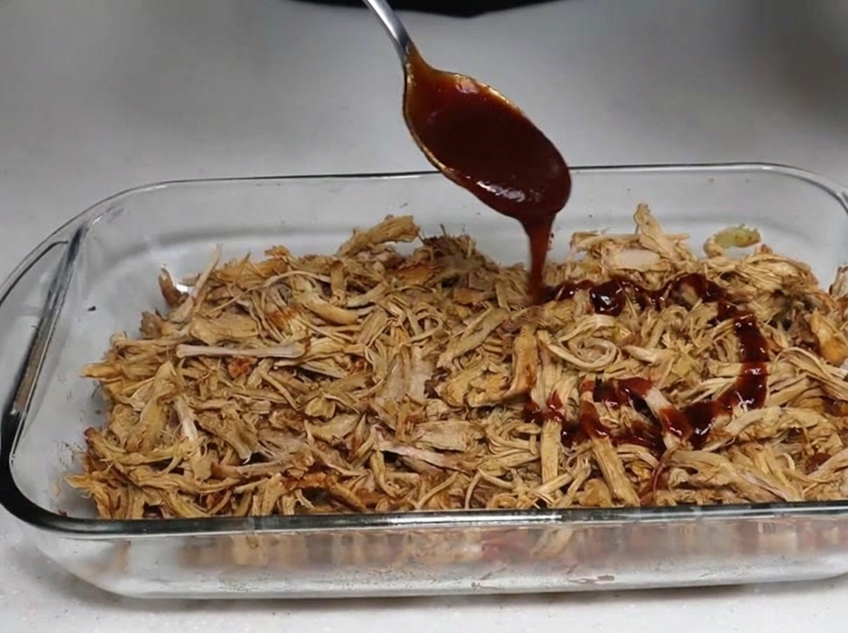 Salsa BBQ versata con un cucchiaio su un piatto di tenero maiale sfilacciato affumicato.