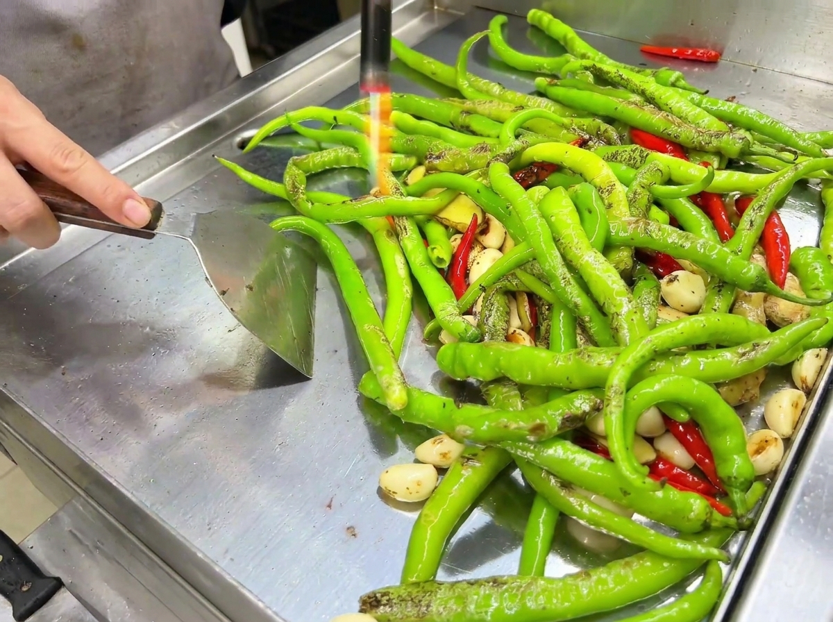 La fiamma di un cannello da cucina che bruciacchia la superficie dei peperoncini sulla piastra.
