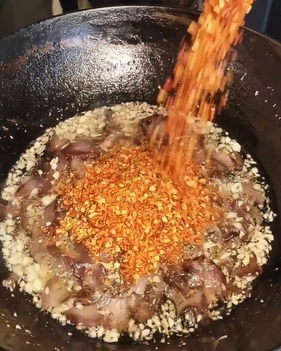 Fiocchi di peperoncino rosso essiccati versati da un contenitore in un wok contenente aglio fritto, carne e olio.
