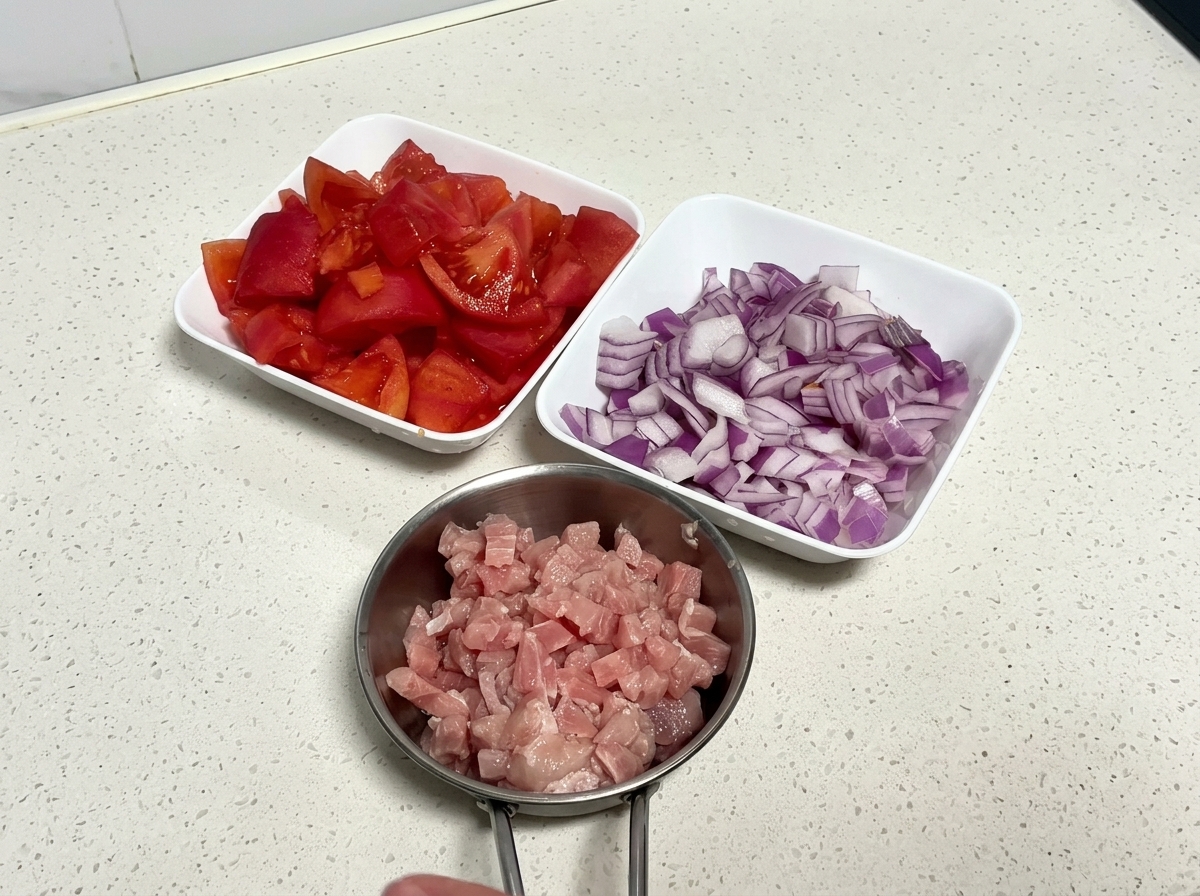 Vista dallalto di tre ciotole bianche contenenti pomodori tritati, cipolla rossa affettata e cubetti di carne di maiale cruda.