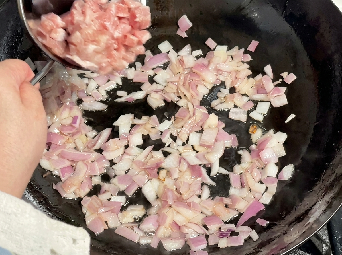 Cubetti di carne cruda versati da una ciotola di metallo in un wok con cipolla soffritta.