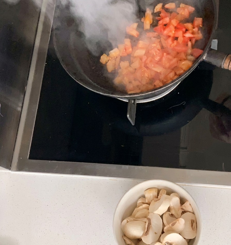 Pomodori a cubetti saltati in una padella nera con una ciotola di funghi affettati sullo sfondo.