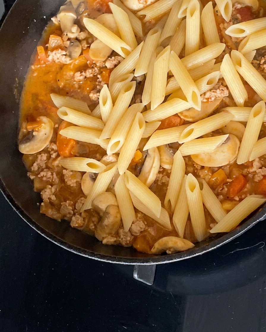 Penne cotte aggiunte al sugo di carne e funghi in padella.