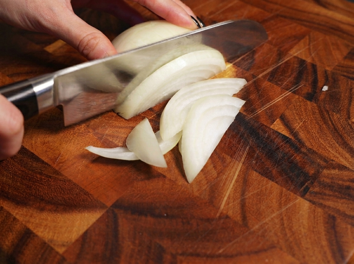 Una mano usa un coltello da chef per affettare una cipolla bianca su un tagliere.