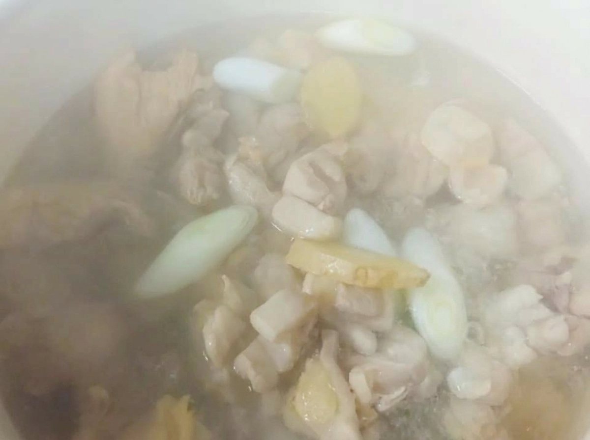 Acqua calda fumante in una pentola bianca sopra i pezzi di pollo, zenzero a fette e cipollotti.