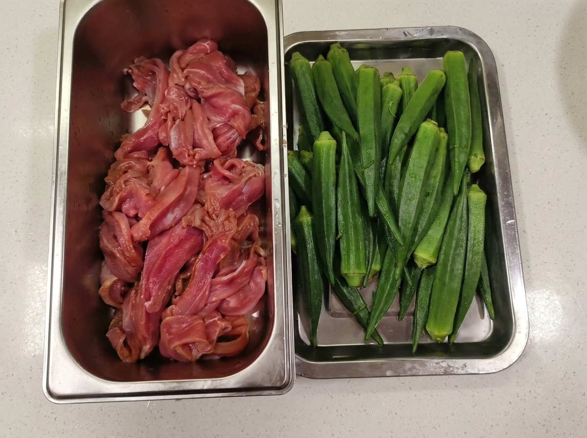 Un contenitore di metallo con carne danatra affettata sottile, posizionato accanto a un vassoio pieno di gombo verde lavato.