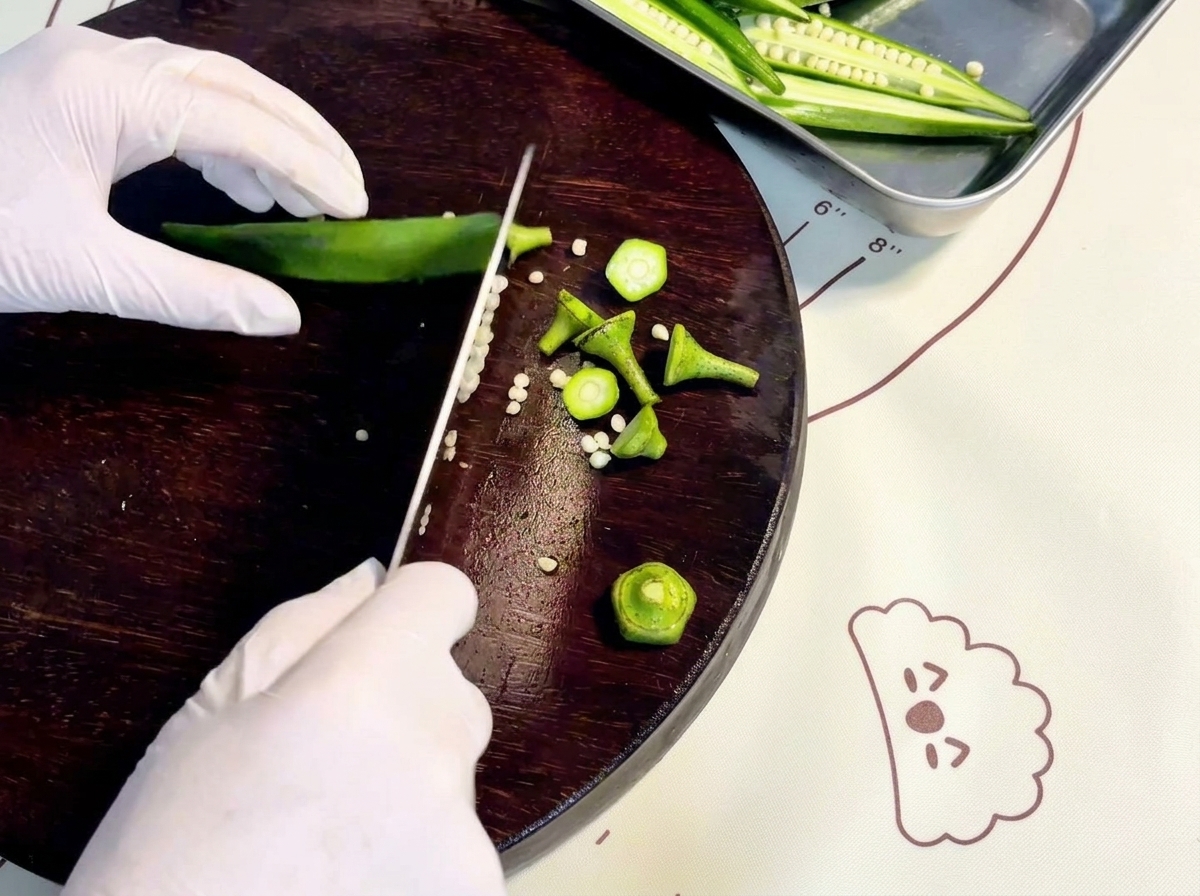 Mani guantate che tagliano okra fresca verde per il lungo su un tagliere di legno scuro.