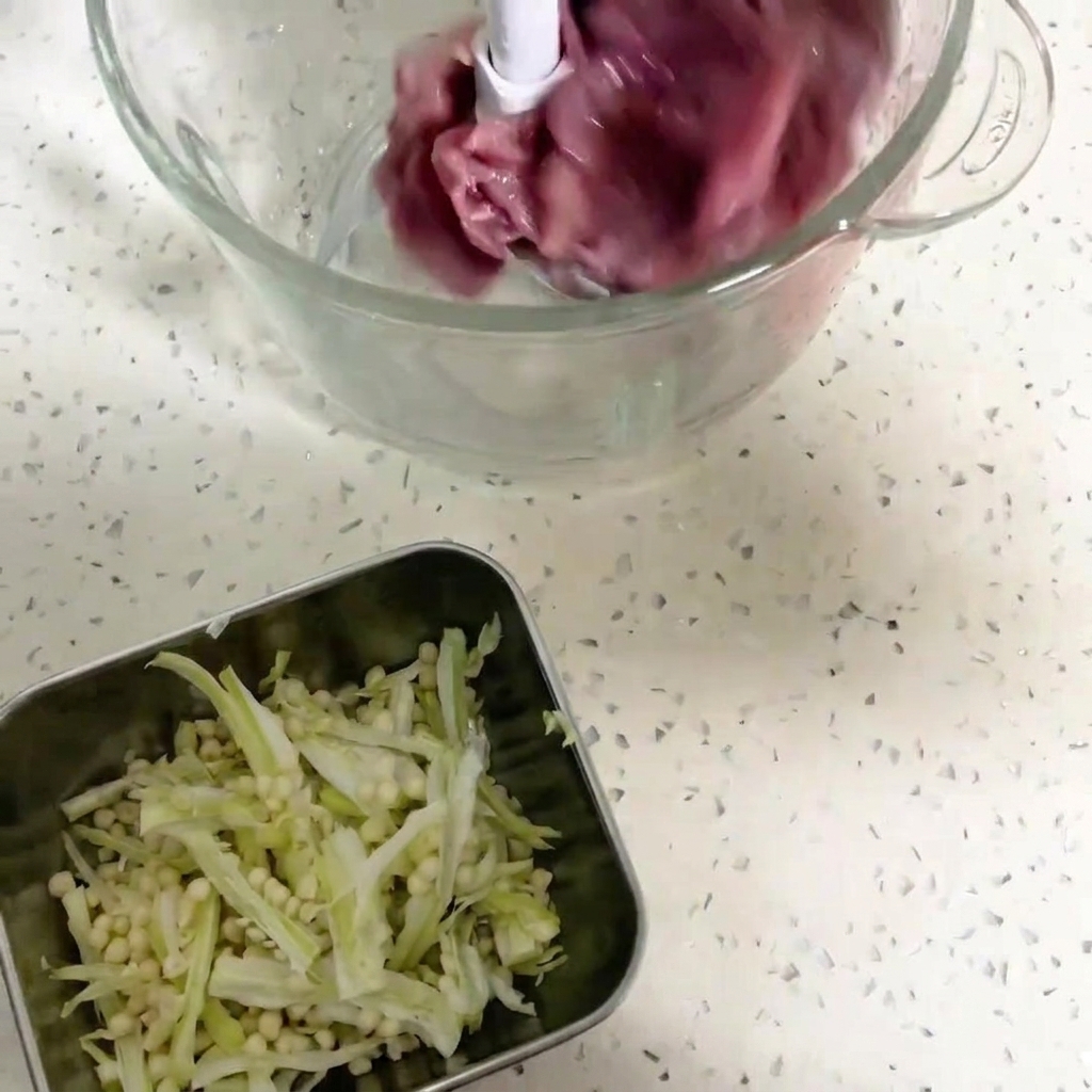 Primo piano della ciotola di un robot da cucina contenente pezzi di carne danatra rossa scura e semi di okra, pronti per essere frullati.