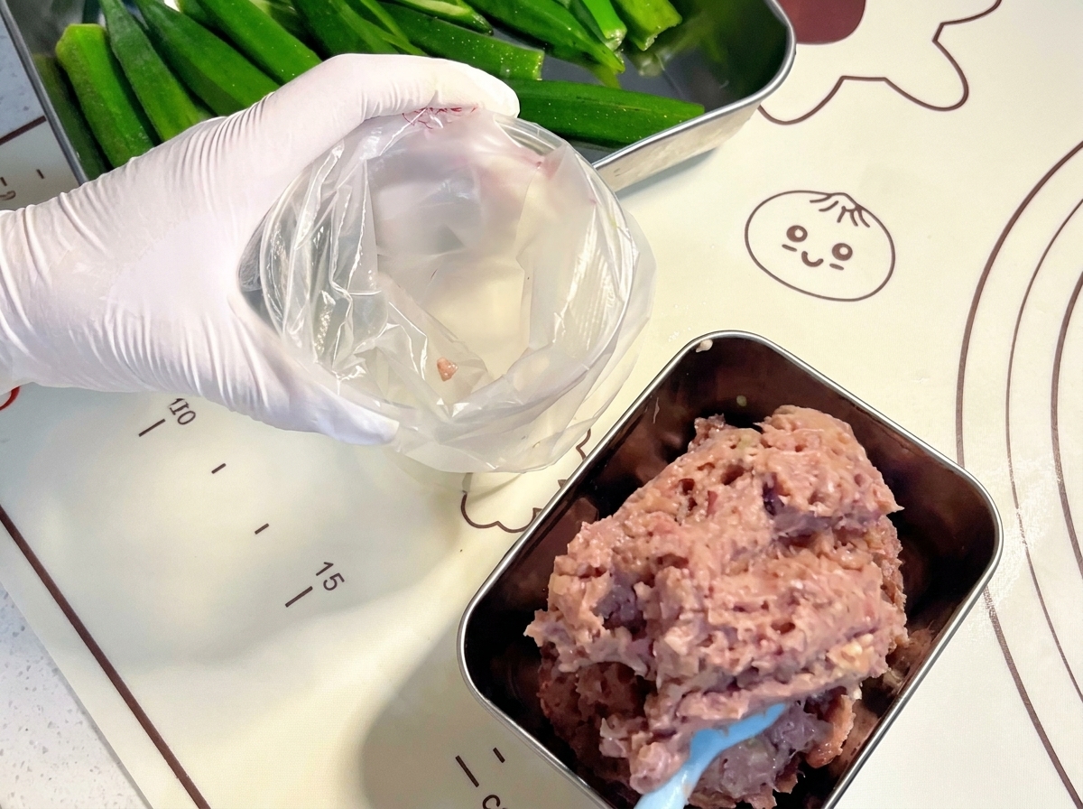 Mani guantate che usano una spatola blu per trasferire la pasta di carne cruda in un sac-à-poche di plastica trasparente.