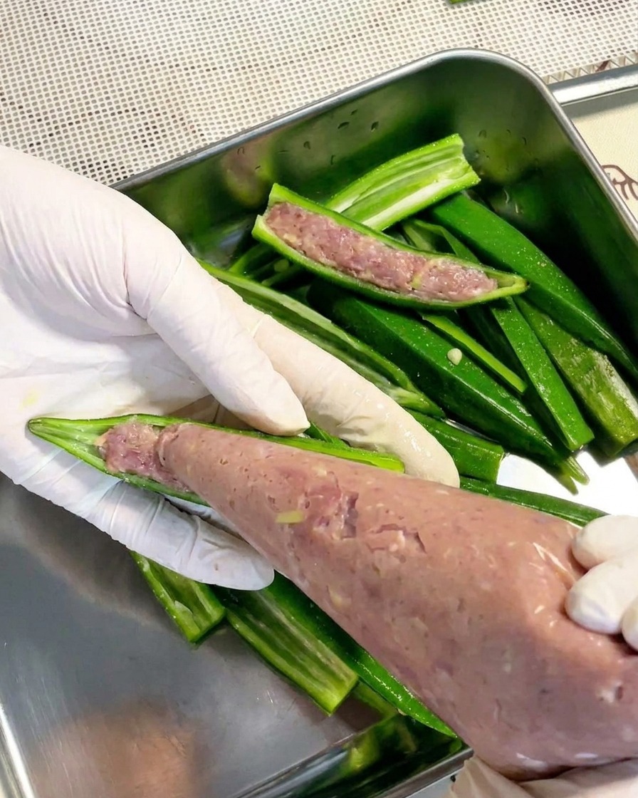 Un paio di mani guantate che usano un sac-à-poche trasparente per riempire una metà di okra sbollentata con una pasta di carne liscia.