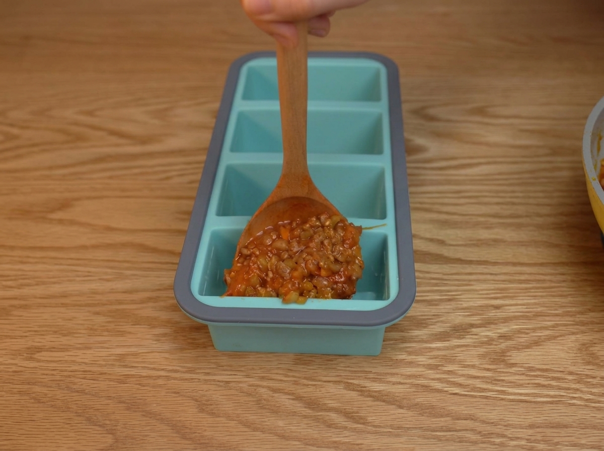 Denso sugo di pomodoro e carne distribuito con un cucchiaio nei singoli scomparti di un vassoio in silicone blu.