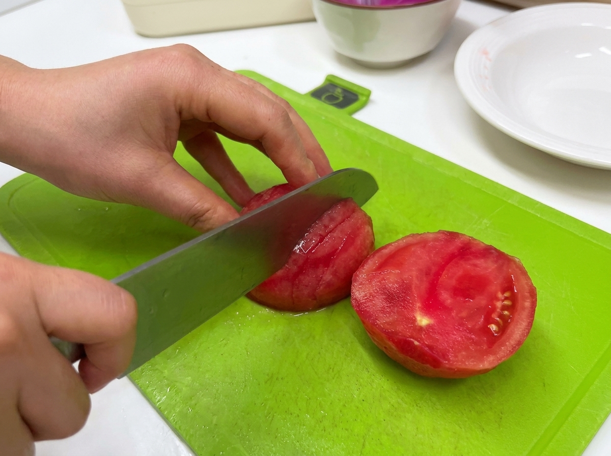 Una persona taglia un pomodoro sbucciato con un grosso coltello su un tagliere verde.