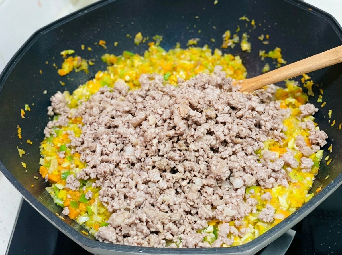 Carne macinata mescolata al soffritto di carote, sedano e cipolle in un grande wok.