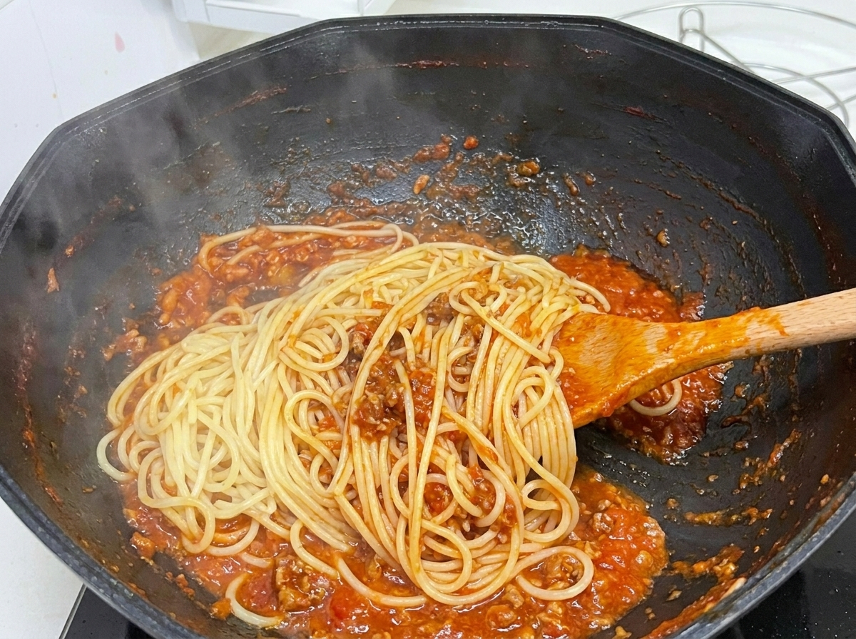 Spaghetti lessati mescolati in un wok nero con il ragù utilizzando un cucchiaio di legno, con vapore che sale.