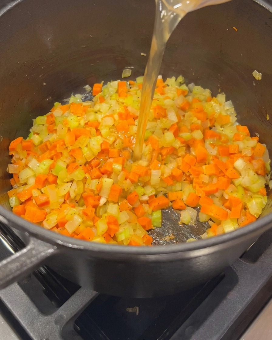 Brodo di pollo dorato versato da un misurino in una pentola con soffritto di verdure.