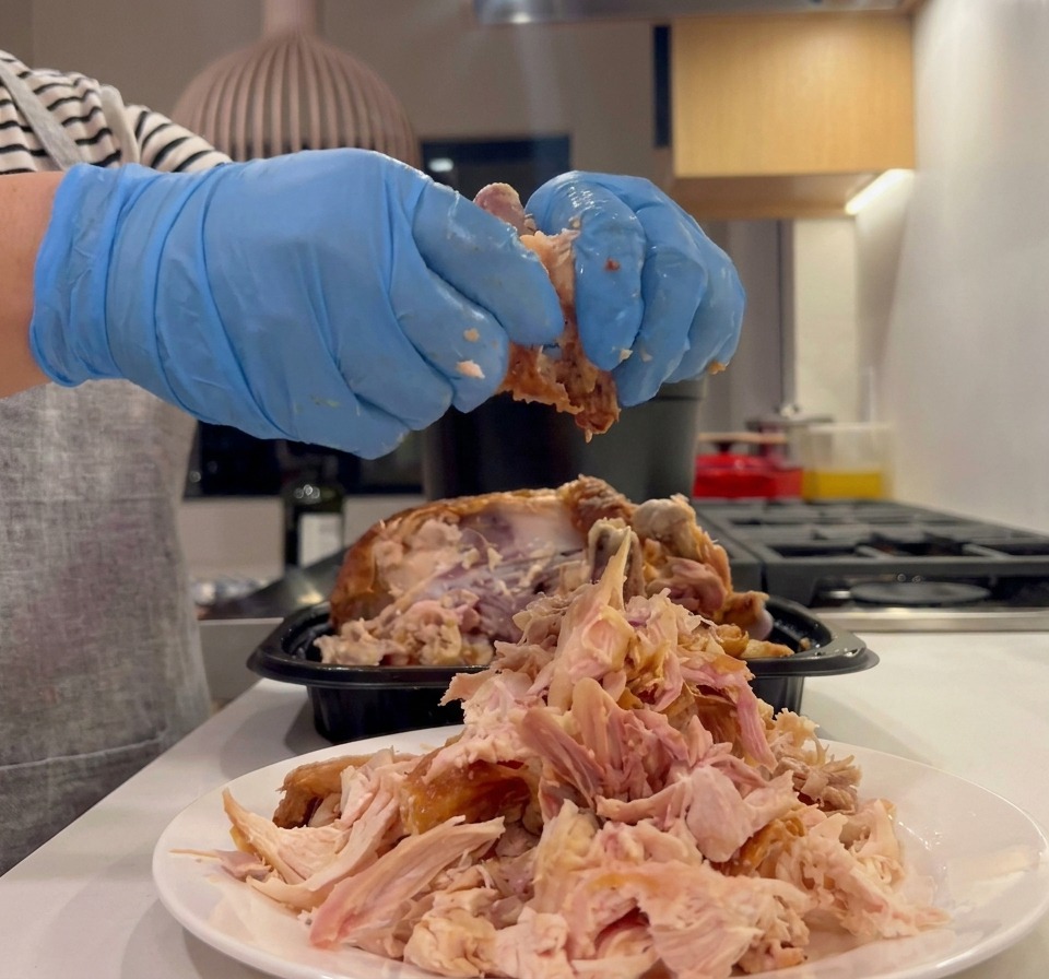 Mani con guanti da cucina blu che sfilacciano pollo arrosto cotto su un piatto bianco.