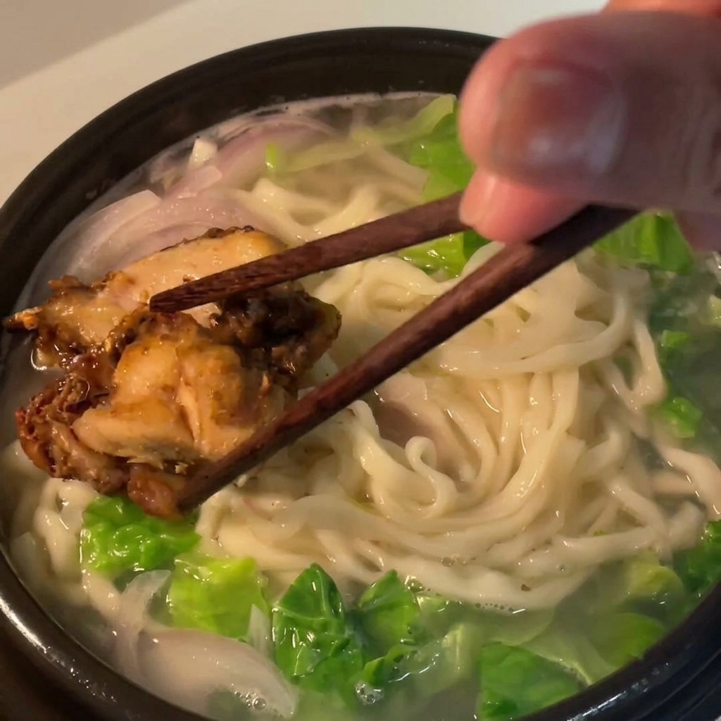 Pezzi di coscia di pollo cotta posizionati sul letto di noodles nella ciotola della zuppa utilizzando bacchette di legno scuro.
