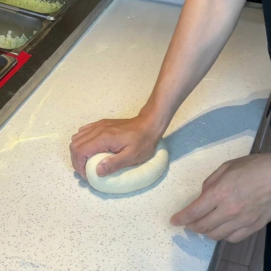 Mani che premono su una pallina di impasto per pizza su un piano di lavoro infarinato.