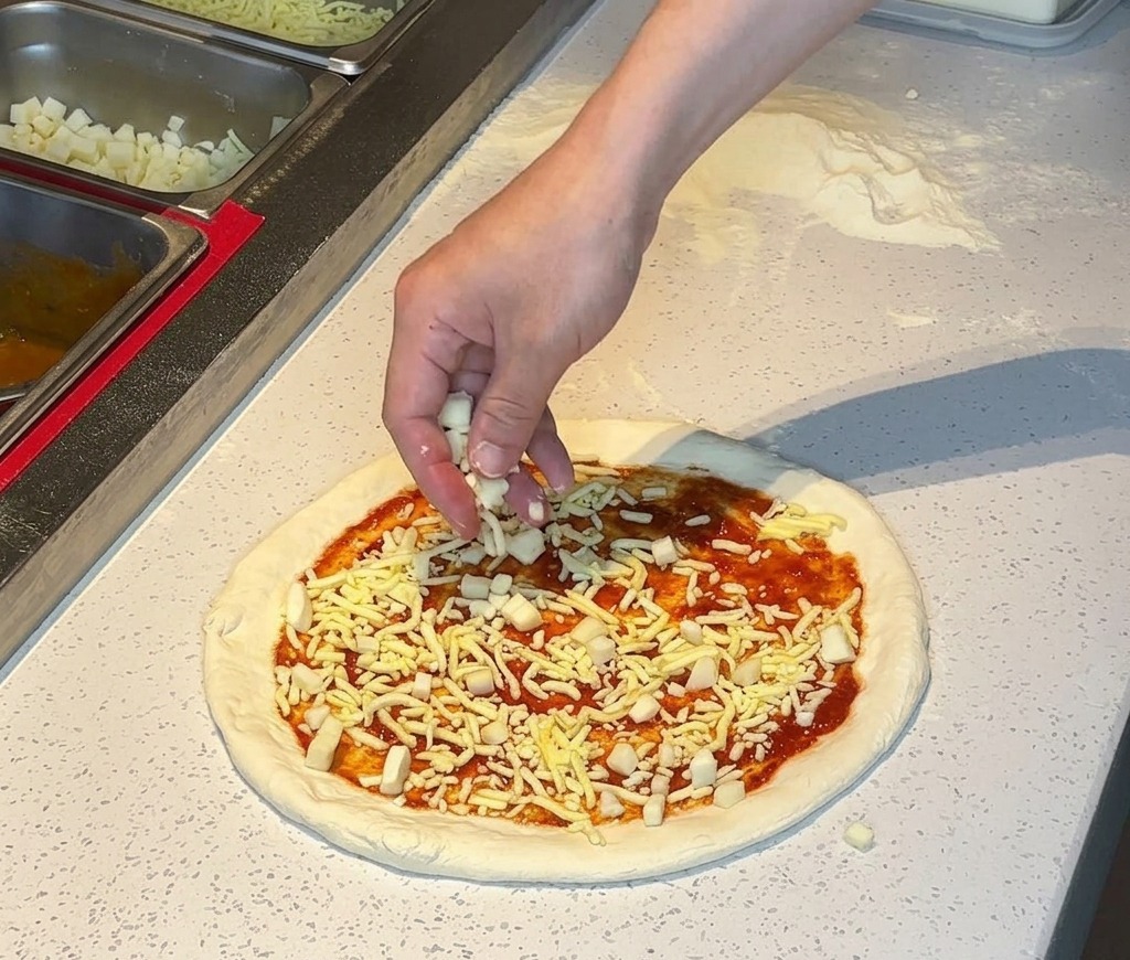 Una mano che sparge mozzarella grattugiata su una base per pizza con salsa.