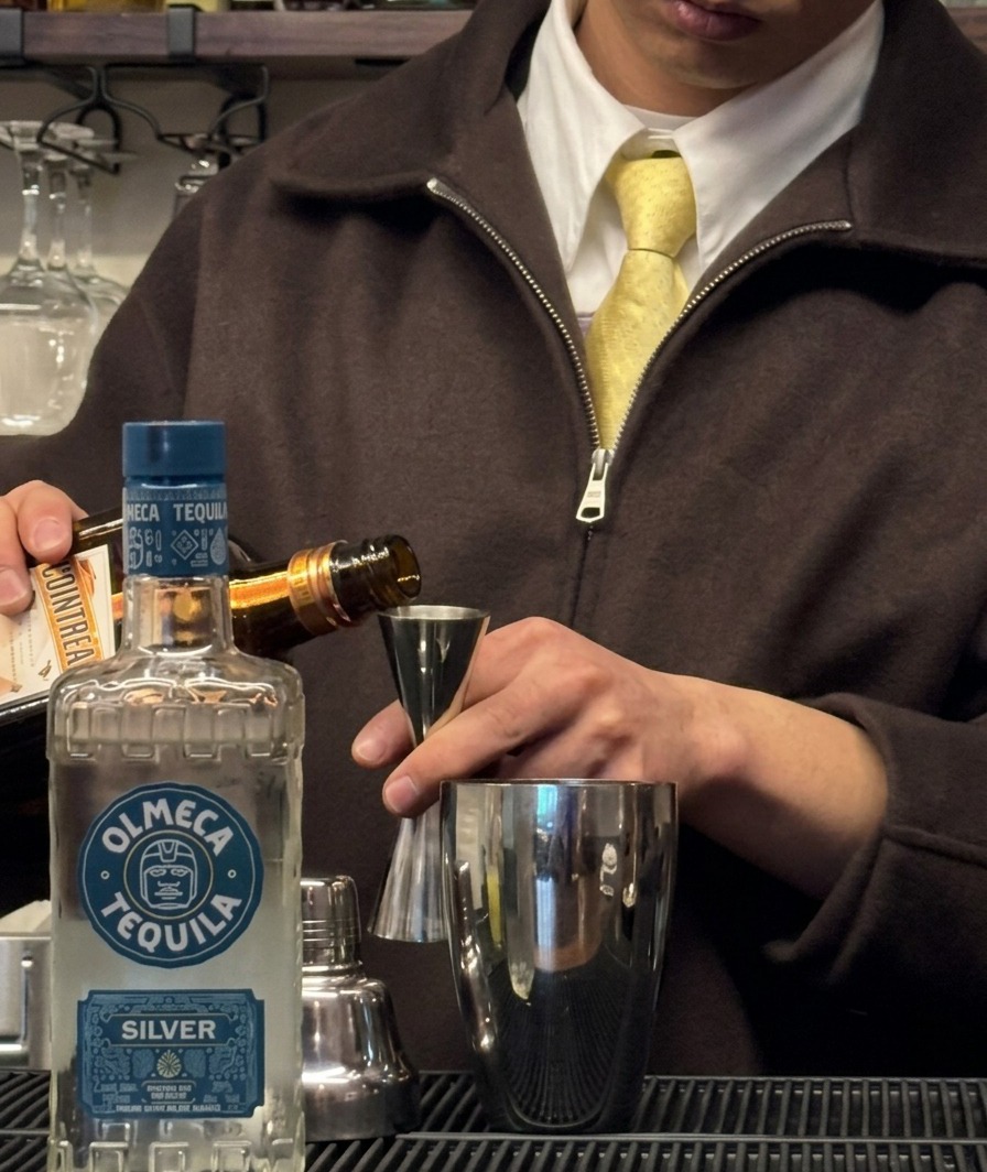 Triple sec versato da una bottiglia in un jigger posizionato sopra uno shaker.