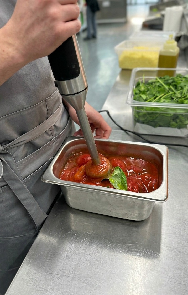 Primo piano di un frullatore a immersione che lavora i pomodori pelati e il basilico per creare una salsa rossa e liscia.