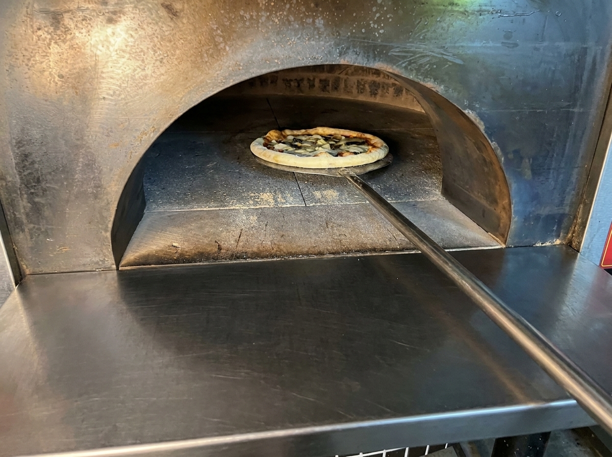 La pizza cruda assemblata viene inserita in un forno a legna molto caldo usando una lunga pala di metallo.