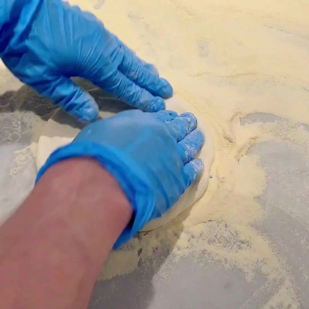 Mani con guanti blu che premono e stendono limpasto crudo per pizza su un piano di lavoro infarinato.