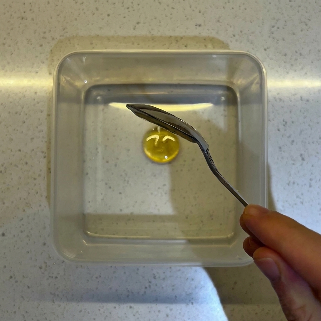 Un primo piano di un cucchiaio che mescola miele e lievito in acqua dentro un contenitore quadrato trasparente.