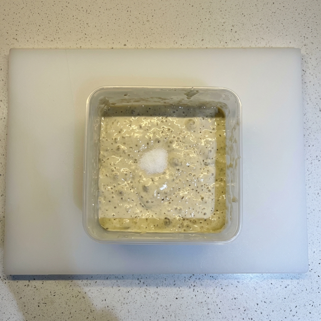 Sale e un mucchietto di farina appoggiati sopra un impasto fermentato e bollicinoso in un contenitore quadrato.