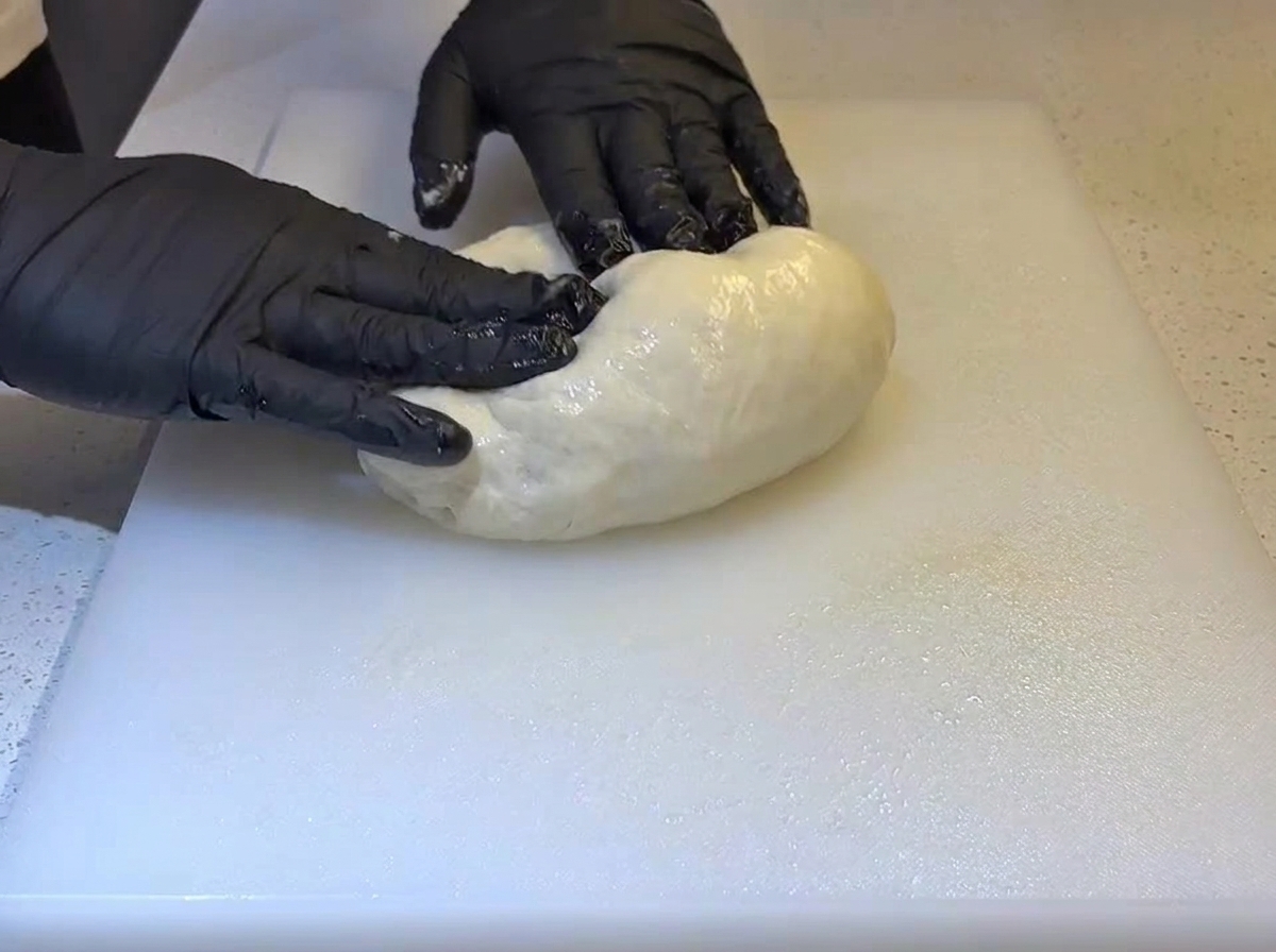 Mani con guanti neri che piegano una palla di impasto morbido e oliato su un tagliere bianco.
