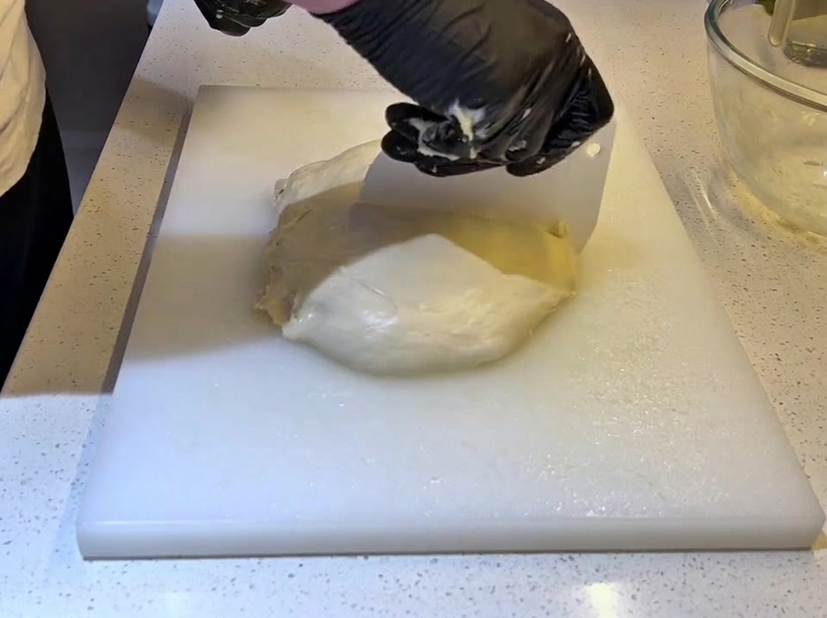Mani con guanti neri che usano un raschietto per impasto in plastica bianca per tagliare un grande pezzo di impasto a metà.