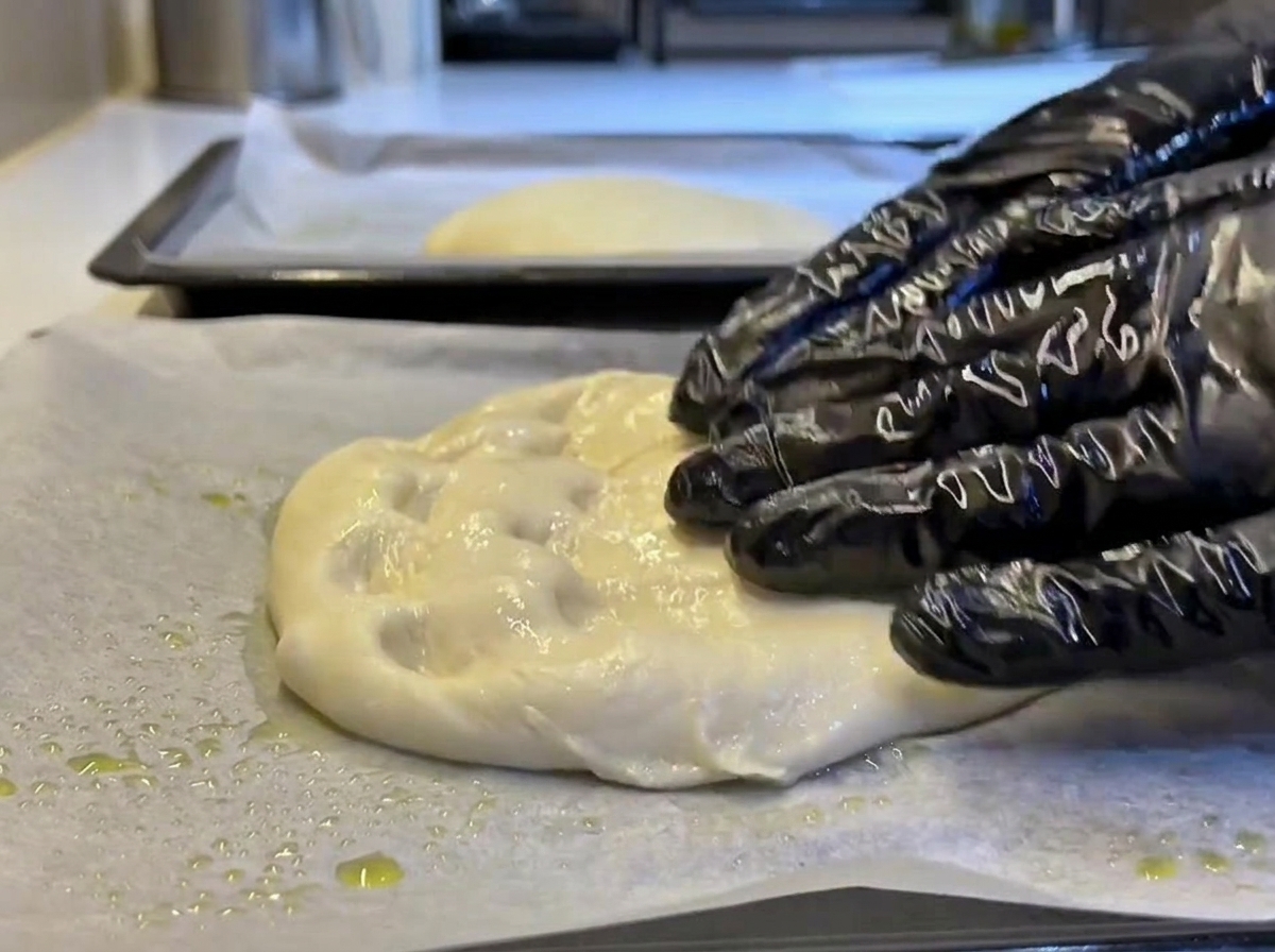 Mani con guanti che premono e stendono delicatamente un pezzo di impasto morbido in un rettangolo su carta forno oliata.