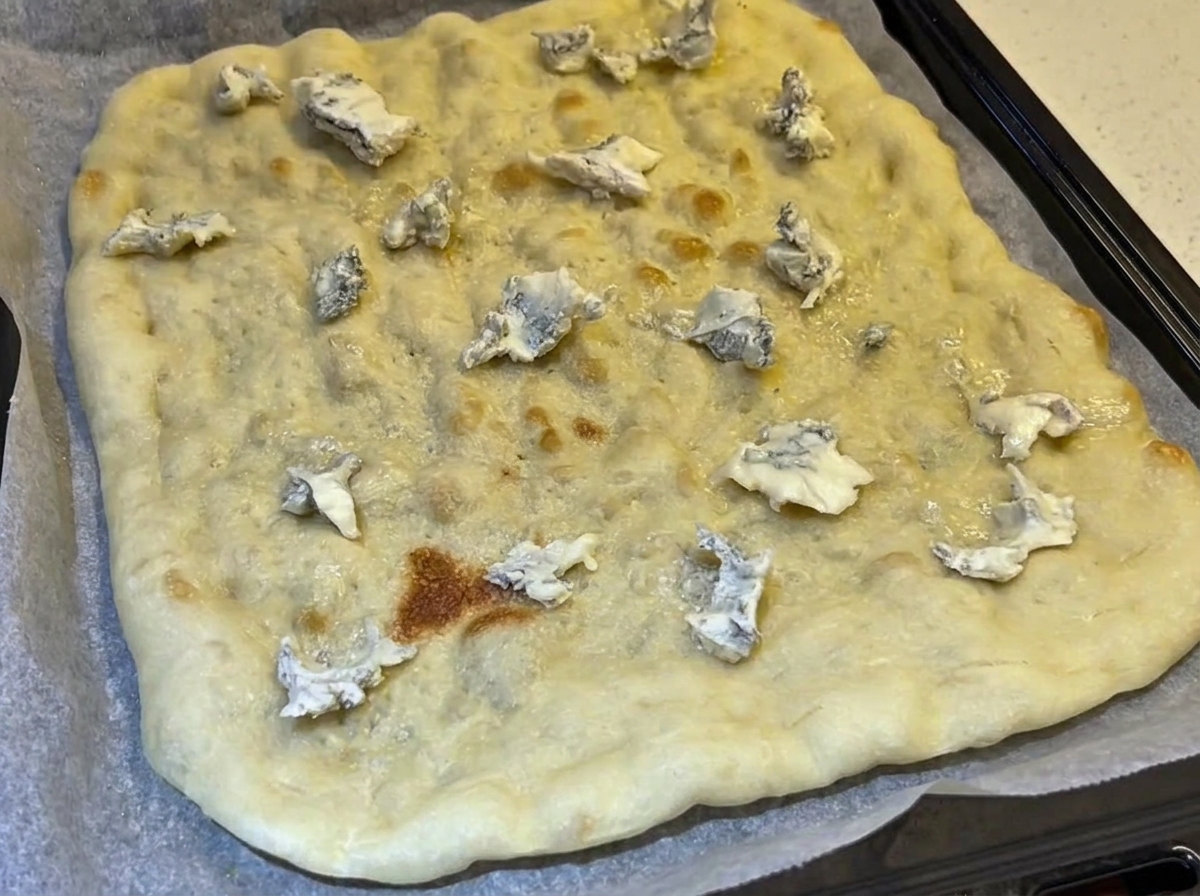Una crosta rettangolare precotta condita con pezzetti sparsi di formaggio blu su una teglia rivestita di carta forno.