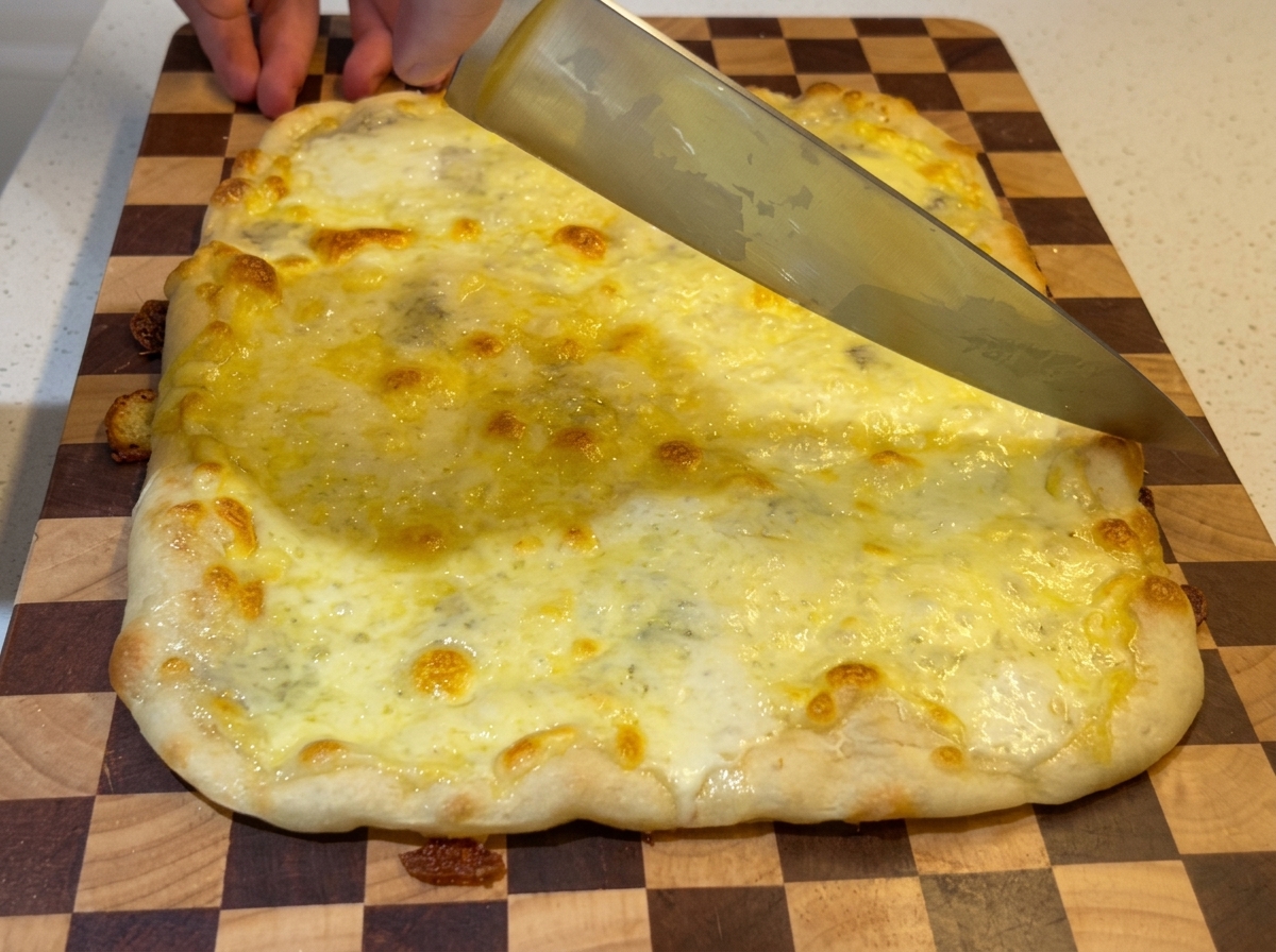 Un grande coltello da chef che taglia una pizza rettangolare ai quattro formaggi fresca e gorgogliante su un tagliere di legno a scacchi.