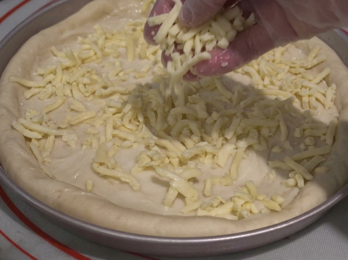 Una mano guantata che sparge abbondante formaggio grattugiato su un impasto per pizza ricoperto di salsa in una teglia.